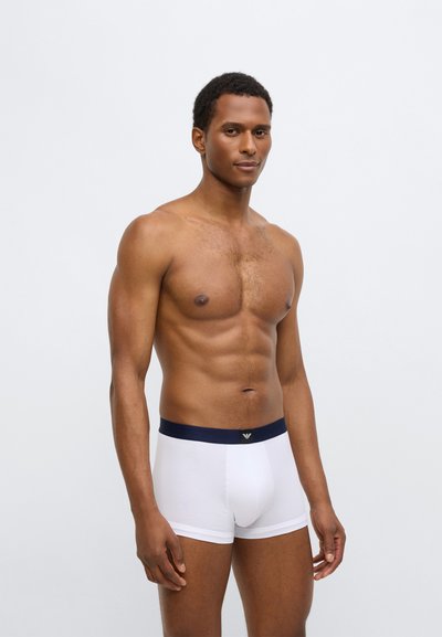 Baltos medvilninės bokserinės trumpikės su tamsiai mėlyna juosmens juosta. Ant juosmens juostos yra logotipas, o suknelė yra prigludusi, pabrėžianti kūno formą.