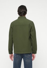 Tumeateeline mees, seljas oliivroheline nööpidega jakk ja sinised teksad, seisab valge ühtlase tausta eest, seljaga kaamera poole.