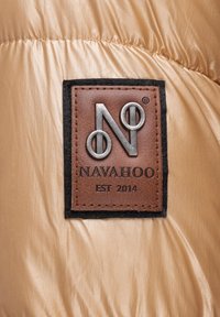 La tela de nylon color beige presenta un parche de cuero marrón oscuro con un logo plateado y el texto "NAVAHOO EST 2014", bordes cosidos y una textura sutil.