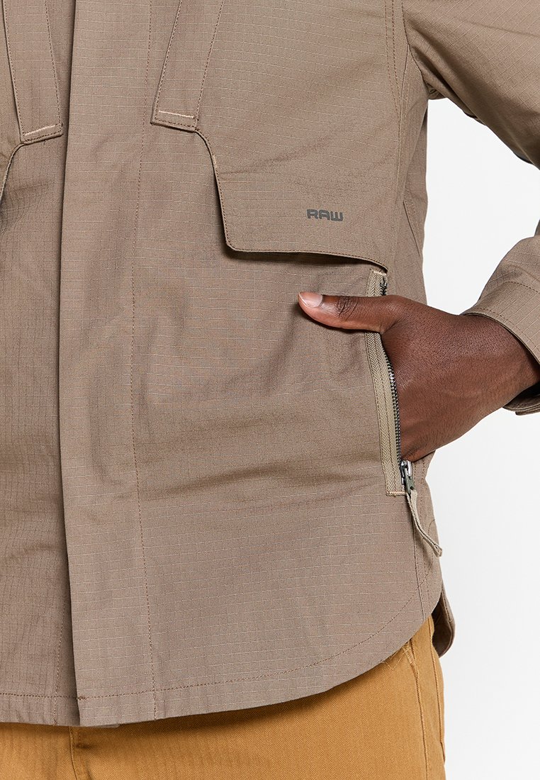 Chaqueta marrón claro hecha de tejido texturizado, con un bolsillo frontal, cierre de cremallera y logo 'RAW'. Presenta un corte relajado y detalles de costura sutiles.