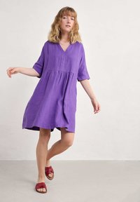 Linen-mekko, joka on violetti, V-pääntie ja lyhyet hihat, jossa on kerätty vyötärö. Yhdistetty punaisiin nauhakenkiin neutraalilla taustalla.