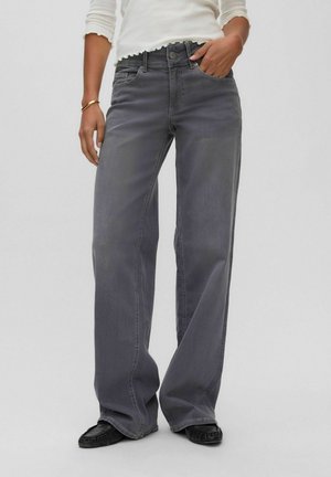 Person iført løstsiddende mørkegrå jeans, hvid langærmet top, sorte sko og et gyldent armbånd med den ene hånd i lommen.