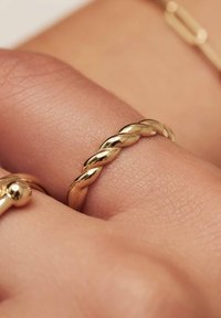 Goldener, verdrehter Ring an einer Hand, der eine glatte, glänzende Oberfläche mit abgerundeten Kanten aufweist. Die Hautstruktur ist im Hintergrund sichtbar.