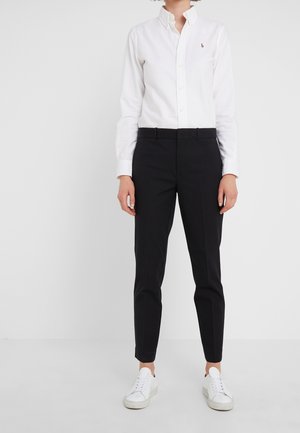 Personne portant une chemise blanche à boutons, un pantalon noir ajusté et des baskets blanches, debout devant un fond clair uni.