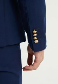 Veste de costume navy avec des poignets ornés de cinq boutons dorés, accompagnés de coutures décoratives à côté des boutons ; texture de tissu lisse.
