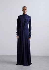 Long navy dress in soft fabric with a high collar, draped details, and long sleeves. Fitted at the waist and flowing to the floor.