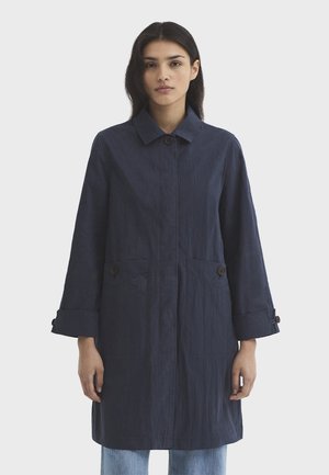 Femme portant un long manteau bleu marine foncé avec un col, des poignets boutonnés, des poches avant et un jean bleu clair, debout devant un fond uni.