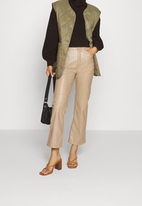 Veste matelassée vert olive, pull marron, pantalon en similicuir beige, et sandales à talons carrés brun clair. Sac à épaule noir complète la tenue.