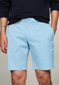 Pantaloni corti di cotone azzurri chiari con fronte piatto, passanti per cintura e due tasche laterali. Un sottile dettaglio del logo ricamato vicino all'orlo.
