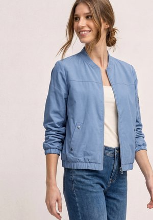 Vrouw glimlacht en kijkt opzij, draagt een lichtblauw bomberjack met elastische manchetten, een wit topje en blauwe spijkerbroek tegen een effen achtergrond.