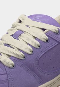 Nærbillede af lilla snøresko i ruskind med hvide teksturerede snørebånd, der er snøret gennem seks snørehuller på hver side.