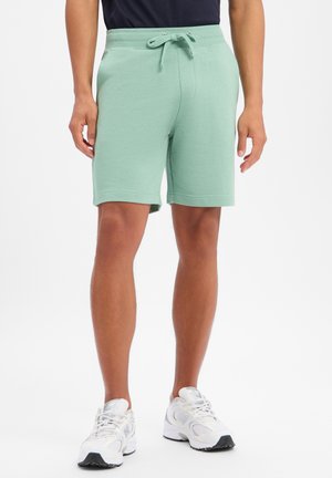 Mężczyzna w jasnoszortach na sznurki i białych butach sportowych, stojący na jednolitym jasnym tle.