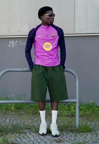 Lilla Nike trøje med guld Spotify-logo, sorte ærmer, grønne shorts, hvide sokker og grå sneakers på en person, der står ved et rækværk.