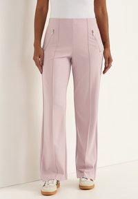 Frau trägt hoch taillierte, hellrosa Hose mit vertikalen vorderen Nähten und diagonalen Reißverschlusstaschen, kombiniert mit weißen Turnschuhen.