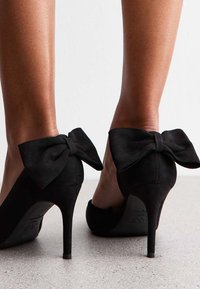 Svarta mocka högklackade skor med en stor rosett bak, med spetsig tå och smal stilettklack. Slät textur.
