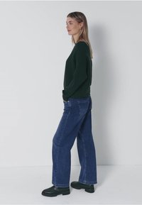 Grüner Strickpullover, blaue weitgeschnittene Jeans und schwarze Knöchelstiefel. Der Pullover hat lange Ärmel; die Jeans verfügen über ein klassisches Fünf-Taschen-Design.