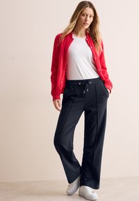 Rote Bomberjacke mit Druckknöpfen, weißem T-Shirt und schwarzen, weit geschnittenen Hosen mit Kordelzugbund. Weiße Sneaker runden das Outfit ab.