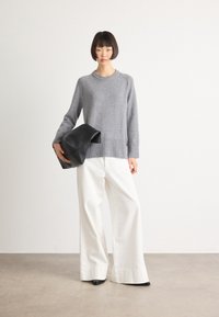 Junge Frau steht auf dem grauen Boden, trägt einen grauen Pullover, weite weiße Hosen, schwarze Schuhe und hält eine große schwarze Clutch vor einem weißen Wandhintergrund.