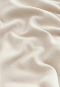 Pehmeä beige kangas hienolla neulepinnalla, joka näyttää hienot laskokset ja varjot luoden sileän, aaltoilevan pinnan.