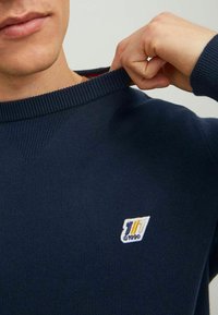 Marineblauer Strickpullover mit geripptem Halsausschnitt, versehen mit einem kleinen weißen gestickten Logo mit gelben und orangen Akzenten auf der Brust.