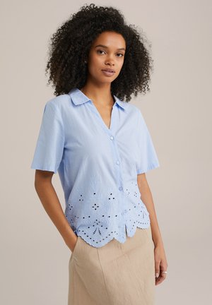 Blusa azzurro chiaro a maniche corte con chiusura a bottoni e motivo floreale ricamato sull'orlo. Texture liscia e dettaglio a bordino scallop.