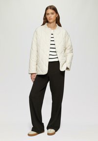 Giacca crema trapuntata con chiusura a scatto, indossata sopra un maglione a righe bianche e nere, abbinata a pantaloni neri a gamba larga e sneakers beige.