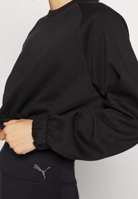 Černá mikina se zaobleným výstřihem a dlouhými, nařasenými rukávy. Obsahuje jemně vyšívané logo a hladkou, měkkou texturu látky.