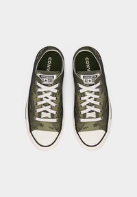 Ζευγάρι Converse All Star χαμηλά σε πράσινο καμουφλάζ με λευκά κορδόνια και καπάκια από καουτσούκ στη μύτη, φωτογραφημένα από πάνω.