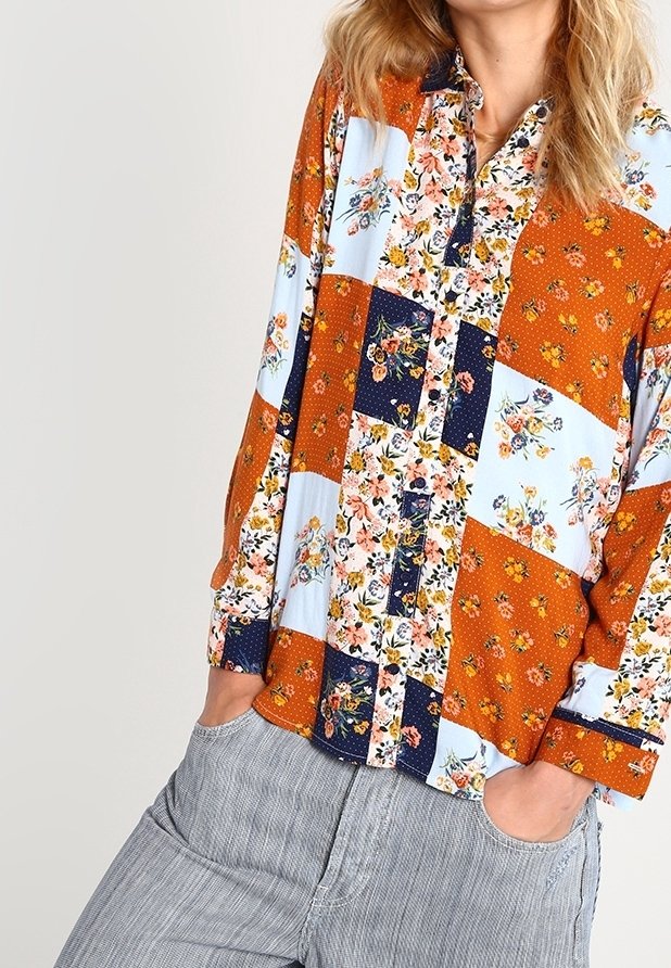Chemise à boutons en patchwork avec des motifs floraux dans des sections orange, bleu marine et bleu clair. Manches longues et col classique.