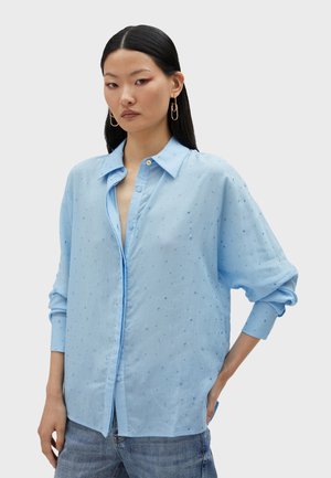 Light blue button-up shirt with a relaxed fit, textured fabric, and small embroidered details. Features a classic collar and rolled sleeves.