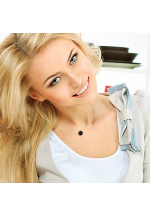 Cheveux blonds, visage souriant, portant une veste beige clair avec un col rayé et un collier délicat avec un pendentif circulaire noir.