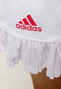Falda atlética blanca con un dobladillo en volantes, que presenta un prominente logo rojo de Adidas en la cintura. Tejido suave con una textura lisa.