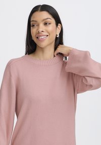 Jasnoróżowy sweter wydziergany, z okrągłym dekoltem ściąganym i luźnymi rękawami, charakteryzujący się miękką fakturą i swobodnym krojem.