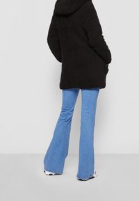 Abrigo negro, esponjoso y con capucha de corte relajado, combinado con vaqueros acampanados de color azul claro y zapatillas blancas. Diseño sencillo, sin patrones visibles.