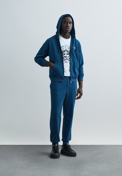 Modrá mikina na zips s pripojenou kapucňou, doplnená o zodpovedajúce tepláky. Tričko má grafický dizajn. Čierne tenisky dokončujú outfit.