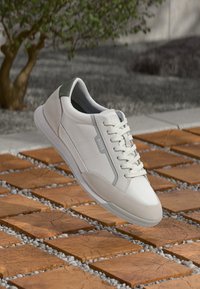White BOSS sneaker with laces and suede accents, hovering above wooden square tiles separated by small gray pebbles outdoors.