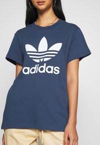 Námorně modré bavlněné tričko s bílým logem trefoil a textem "adidas" ve středu. Krátké rukávy a volný střih.