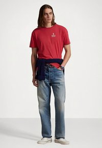 Κόκκινη βαμβακερή μπλούζα με μικρό λογότυπο, συνδυασμένη με ανοιχτόχρωμα τζιν παντελόνια. Ένα σκούρο μπλε πουλόβερ δεμένο γύρω από τη μέση και λευκά αθλητικά παπούτσια ολοκληρώνουν την εμφάνιση.