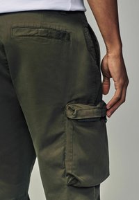 Pantaloni cargo verde măsliniu cu un buzunar lateral cu fermoar, buzunar plat pe spate și textură netedă, evidențiind o croială ajustată și un material durabil.
