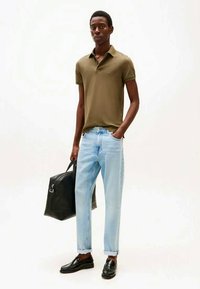 Homem com uma camisa polo verde-oliva, jeans azul claro com as bainhas dobradas, sapatos sociais pretos, segurando uma bolsa de couro preta, de pé contra um fundo branco.
