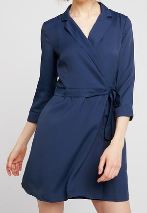 Frau in einem knielangen marineblauen Wickelkleid mit dreiviertel Ärmeln und gebundenem Taillengürtel, die vor einem schlichten Hintergrund steht.