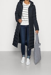 Marineblå puffjakke med hætte og lang længde, parret med en hvid og marineblå stribet top, mørke jeans og hvide sneakers. Stribet tote bag.