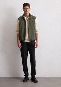 Bezrękawnik w oliwkowej zieleni na beżowej koszuli z długim rękawem, zestawiony z czarnymi spodniami i czarnymi butami. Prosty fason, minimalna ilość okuci.