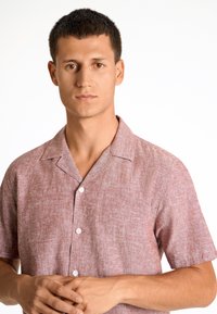 Camisa de manga corta con cuello en un tejido texturizado de color rojo claro y un patrón sutil; cuenta con botones blancos en la parte delantera.
