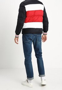 Man med blå uppvikta jeans, vita sneakers och en svart, röd och vit randig puffjacka, ståendes mot en enkel bakgrund.