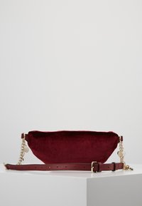 Bolso de terciopelo burdeos con textura suave, con detalle de cadena dorada y correa de cuero ajustable. Forma ovalada y estilizada, diseño minimalista.