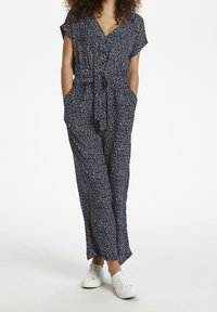 Femme portant une combinaison à motifs floraux bleu marine avec des manches courtes et des baskets blanches, les mains dans les poches, se tenant sur un fond blanc.