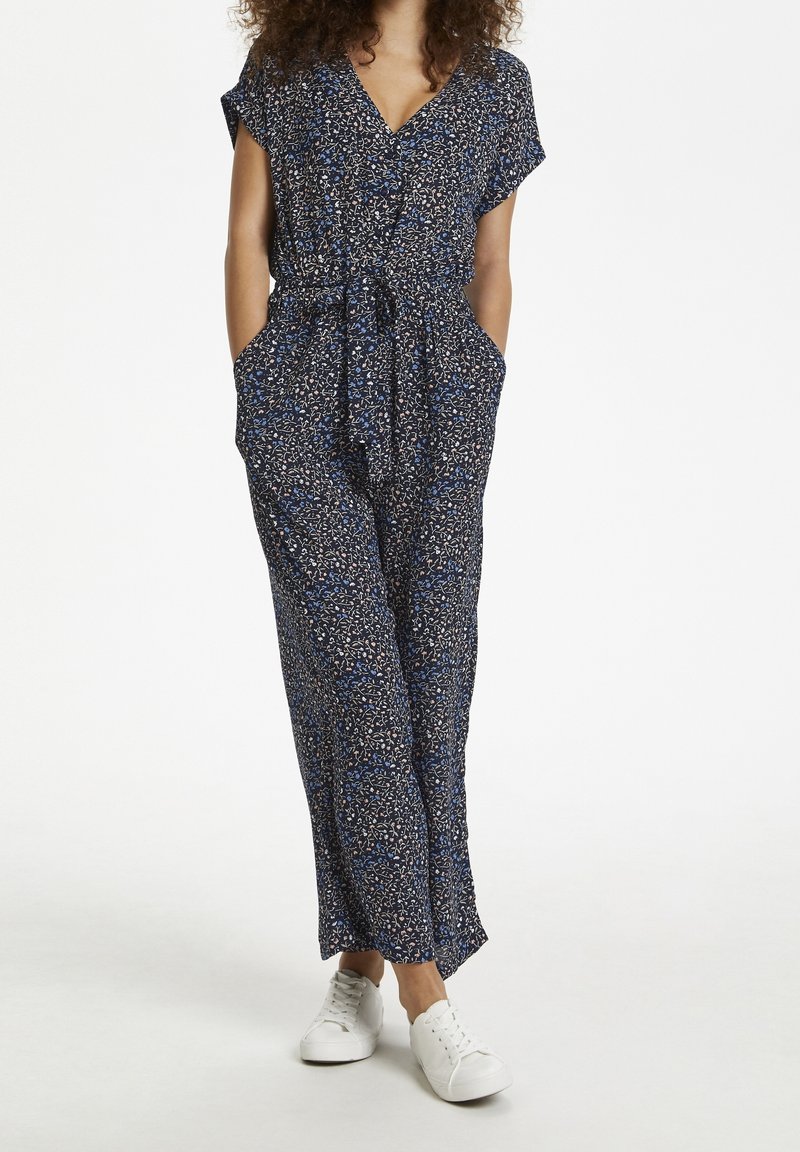 Femme portant une combinaison à motifs floraux bleu marine avec des manches courtes et des baskets blanches, les mains dans les poches, se tenant sur un fond blanc.