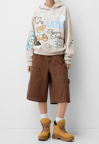 Huvtröja i beige med grafiska tryck, kombinerad med bruna cargo-shorts i knälängd och gula snörboots med kraftig sula.