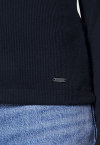 Granatowy sweter z prążkowanego materiału o fakturowym wykończeniu, z małą czarną metką logo na dolnym brzegu, w połączeniu z jasnoniebieskimi dżinsami.